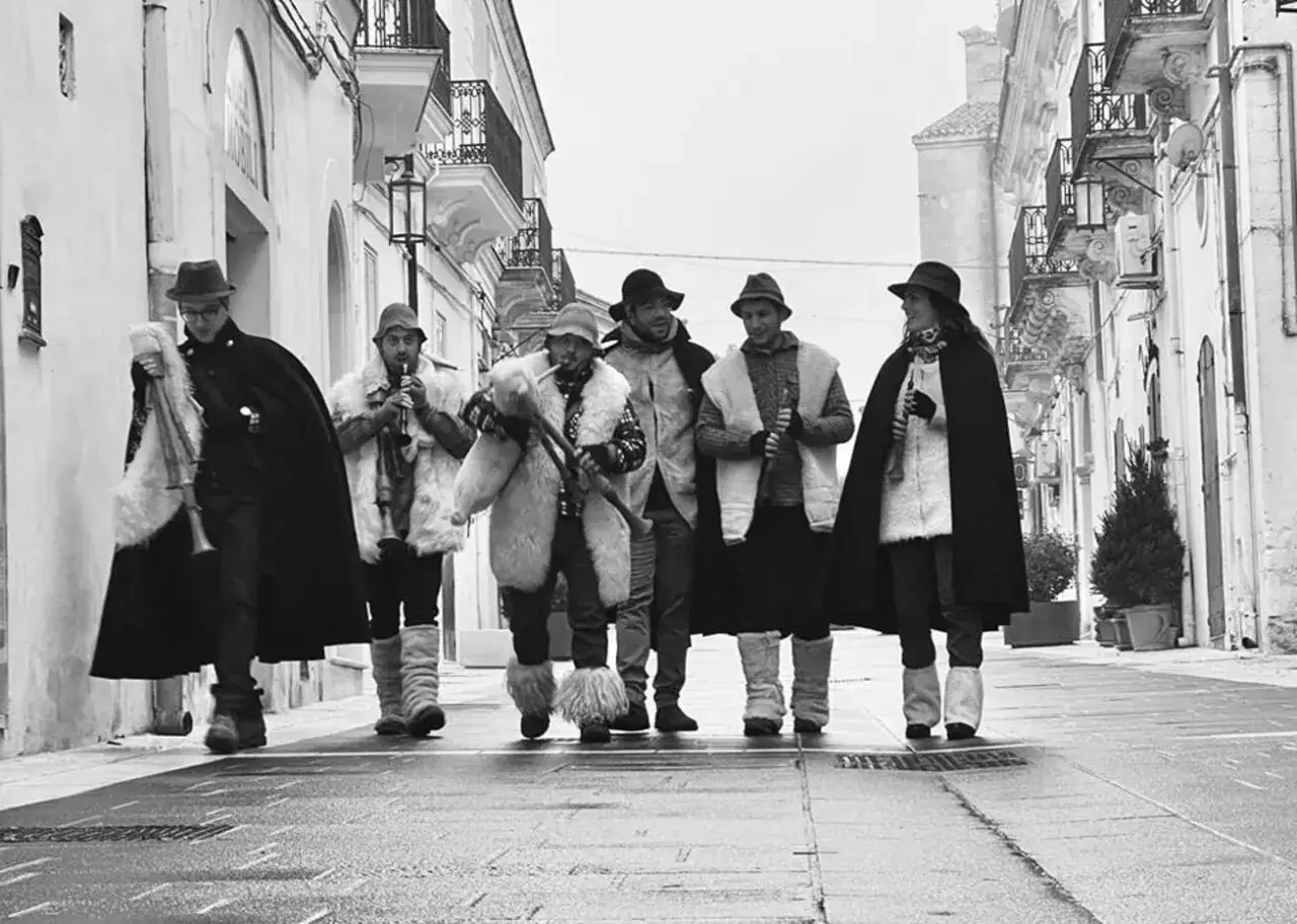 Un gruppo di musicisti con le **piva piva** suona per le strade di un borgo antico, vestiti con abiti tradizionali e cappotti di pelliccia.