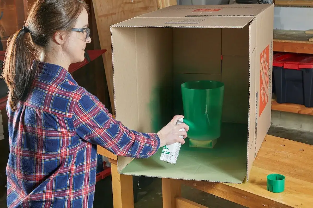 Mujer pintando un jarrón verde con pintura en spray dentro de una caja de cartón. Usa pintura para pintar plástico.