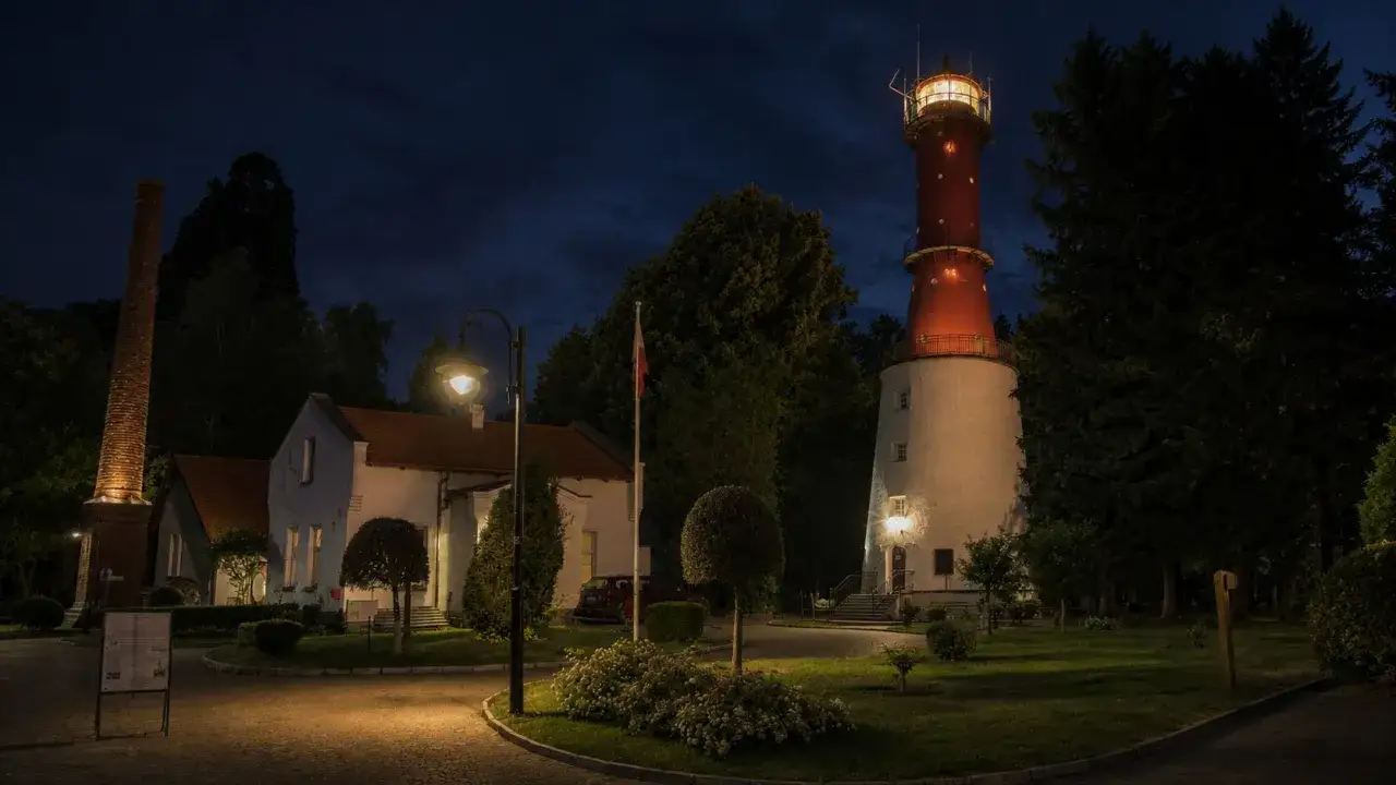 Nocą rozświetlona latarnia morska Jastrzębia Góra strzeże wybrzeża. Obok niej stoją budynki i ceglany komin.