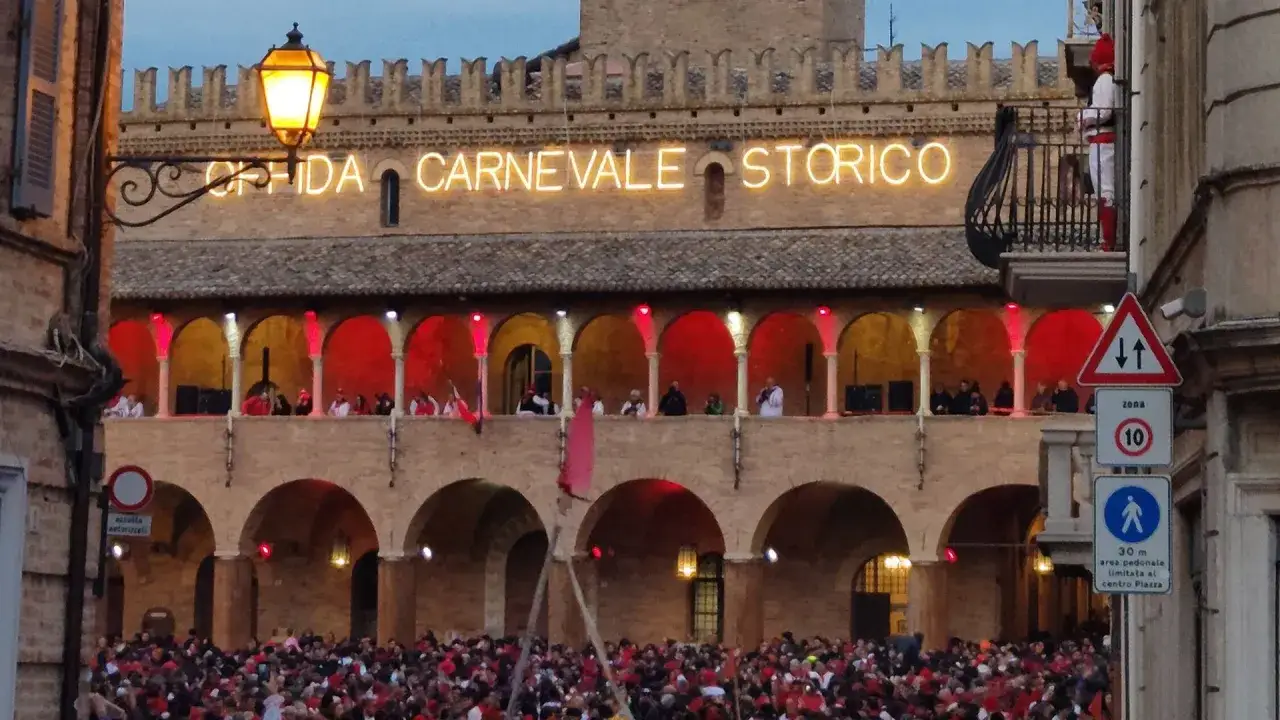 Folla in festa per il Carnevale di Offida. Edificio storico illuminato con scritte al neon e balconata con figuranti in costume.