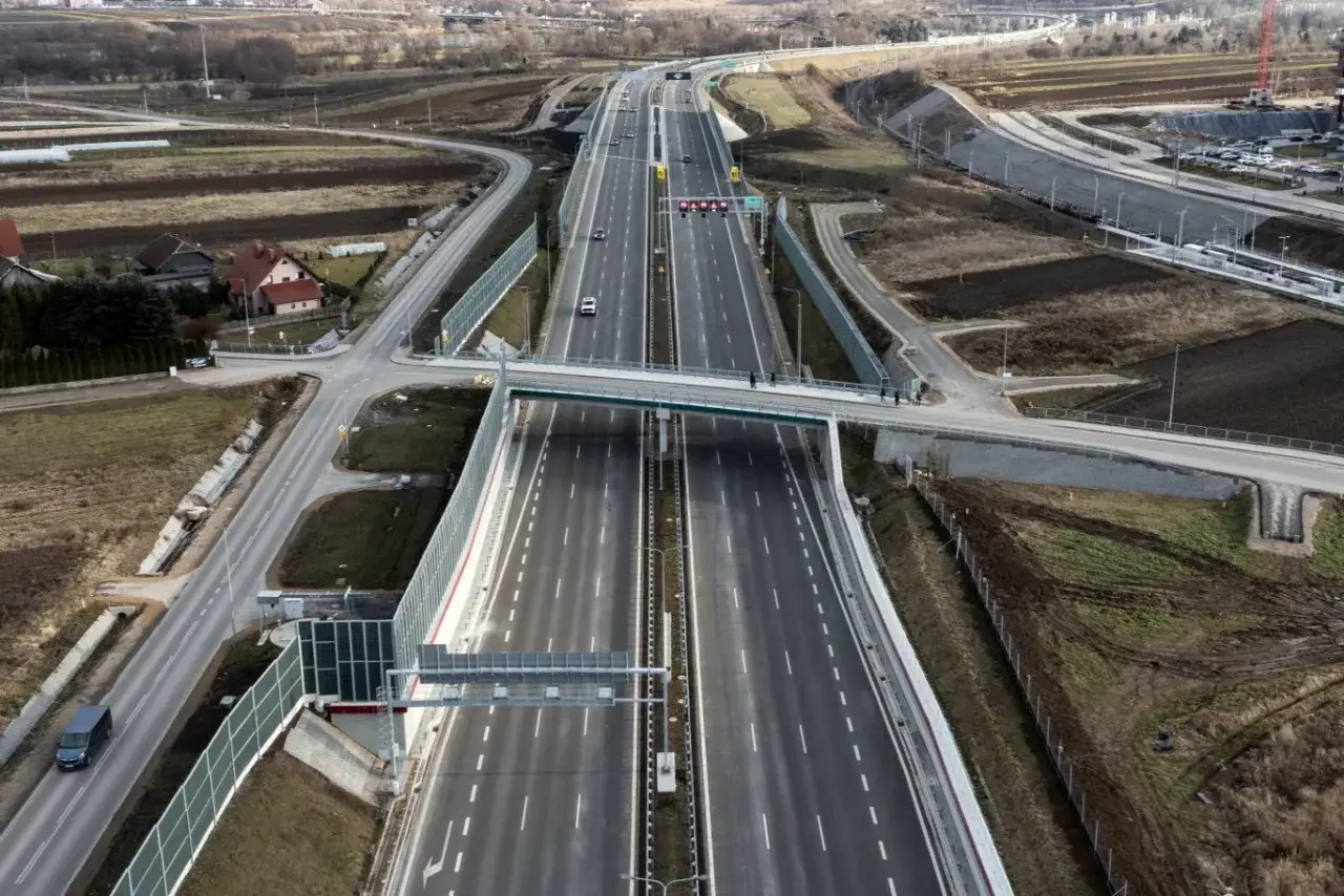 Szeroka autostrada z ruchem pojazdów, z mostem dla pieszych i otaczającym krajobrazem. Prowadzenie pojazdu w tym miejscu jest płynne.