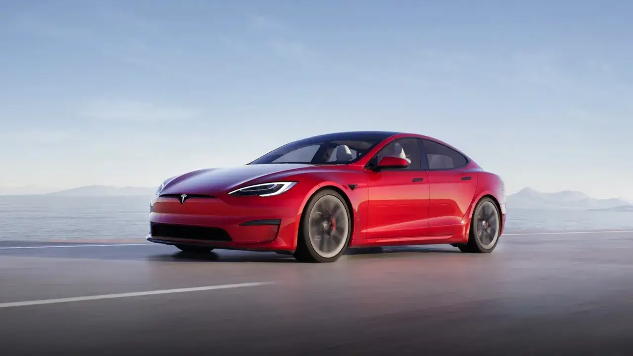 Une Tesla Model S Plaid rouge vif roule sur une route côtière, le soleil se reflétant sur sa carrosserie élégante.