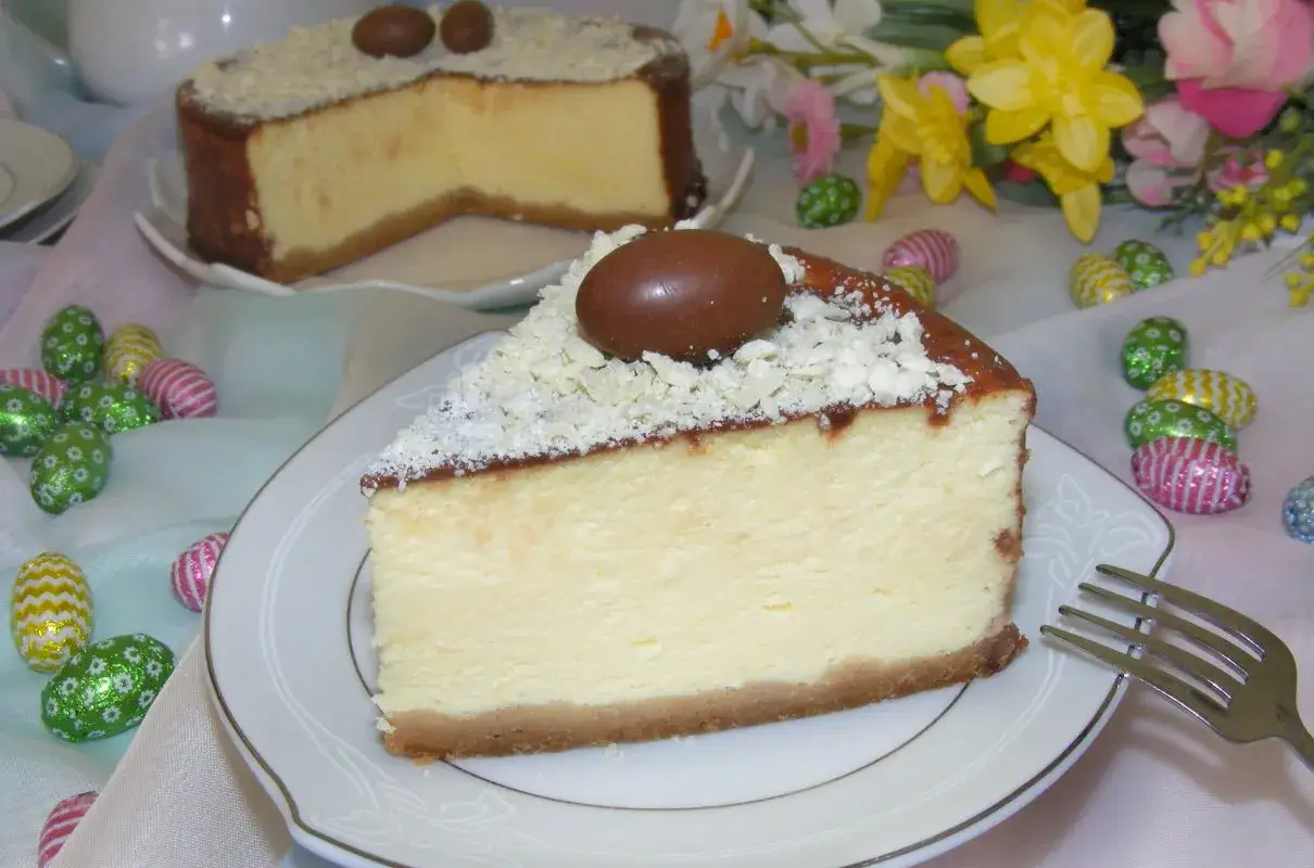 Najdelikatniejszy sernik z mascarpone na zimno, ozdobiony czekoladowymi jajeczkami i wiórkami białej czekolady, na tle kolorowych pisanek.