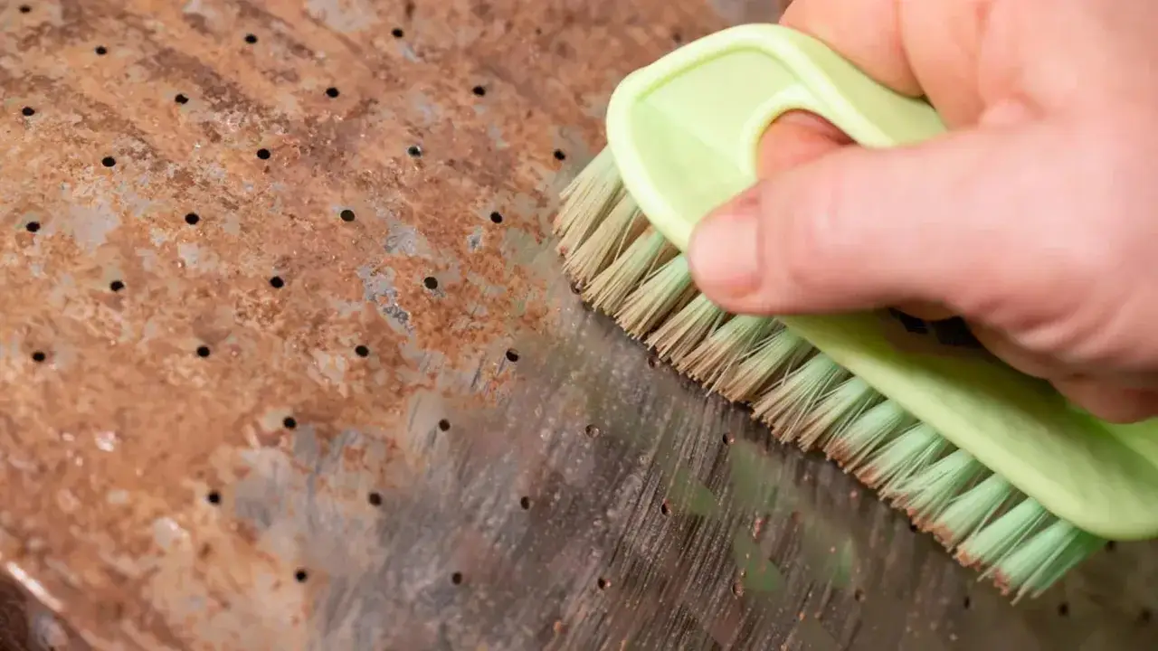 Mano con cepillo verde limpiando una superficie met&aacute;lica oxidada con peque&ntilde;os agujeros.