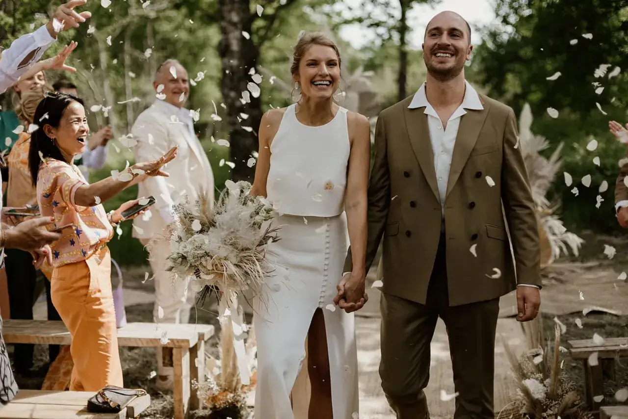 Un couple rayonnant, tel un poème mariage Baudelaire, traverse une pluie de pétales. Le marié en costume vert, la mariée en blanc, entourés de joie.