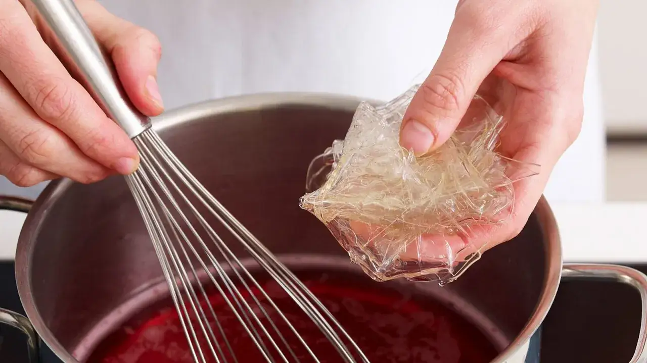 H&auml;nde bereiten Gelatine-Bl&auml;tter f&uuml;r die Zubereitung vor, indem sie sie in eine rote Fl&uuml;ssigkeit in einem Topf geben, w&auml;hrend ein Schneebesen bereitsteht.