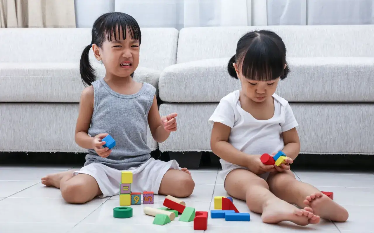 Two young girls engage in parallel play with colorful wooden blocks. One girl looks upset, while the other is focused on her toys.