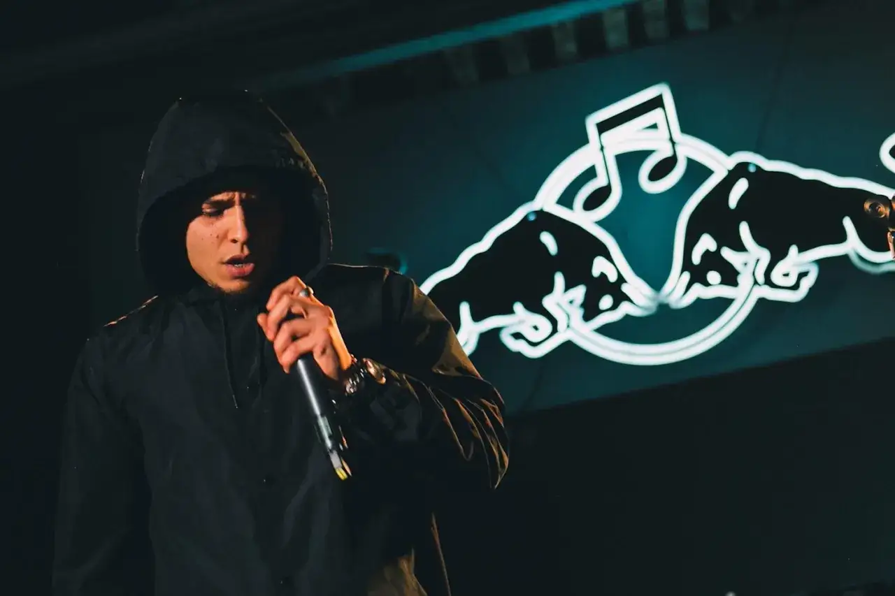 Raper w kapturze, z mikrofonem w dłoni, na tle neonowego logo Red Bulla. Jego słowa niosą ocean wisdom.