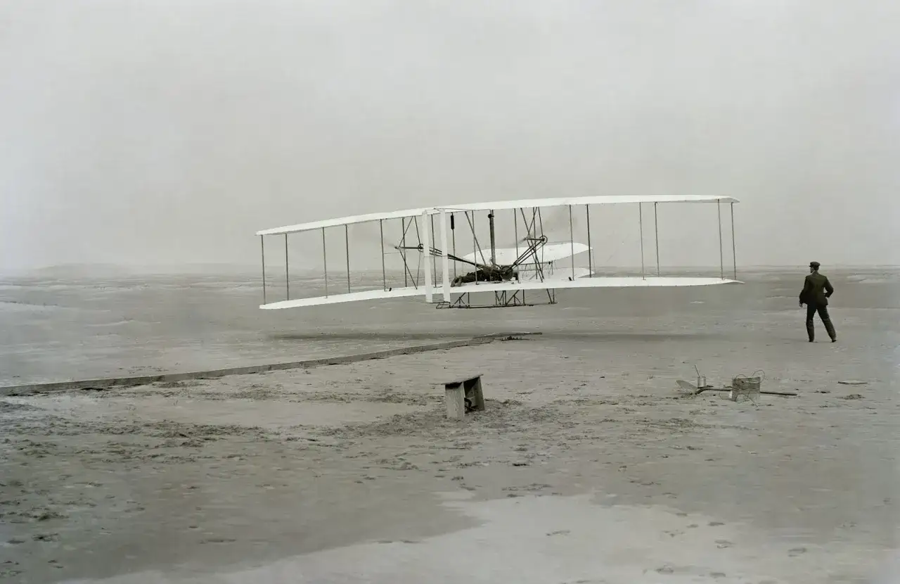 Pierwszy lot samolotem odczucia: Wright Flyer unosi się nad piaszczystym terenem, a jego twórca obserwuje z boku.