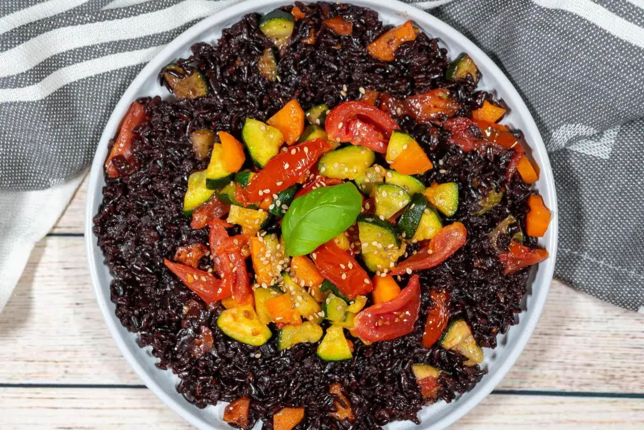 Piatto di riso nero con zucchine, pomodorini e peperoni, guarnito con semi di sesamo e una foglia di basilico. Un'esplosione di colori e sapori.