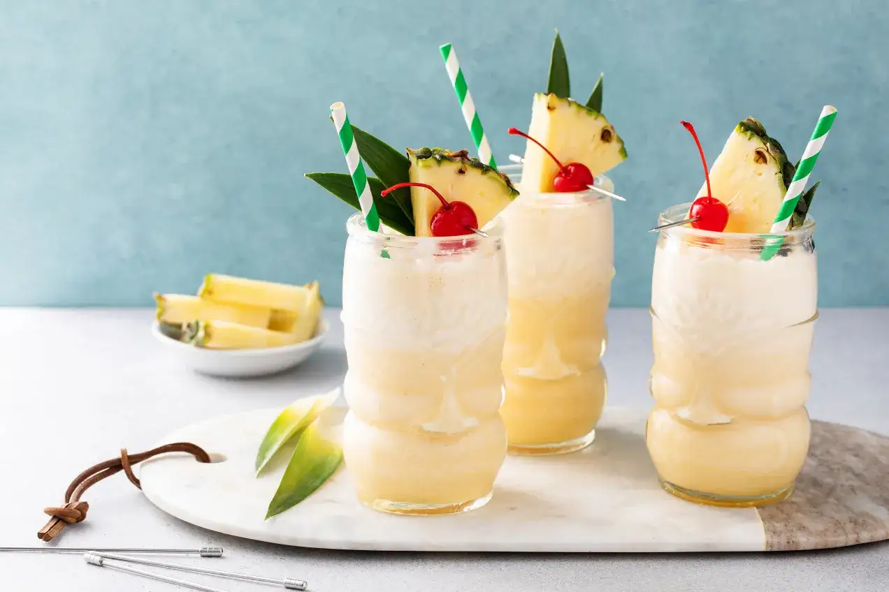 Trzy drinki bezalkoholowe Mocktail w szklankach tiki, ozdobione ananasem i wiśnią. Obok miseczka z kawałkami ananasa.