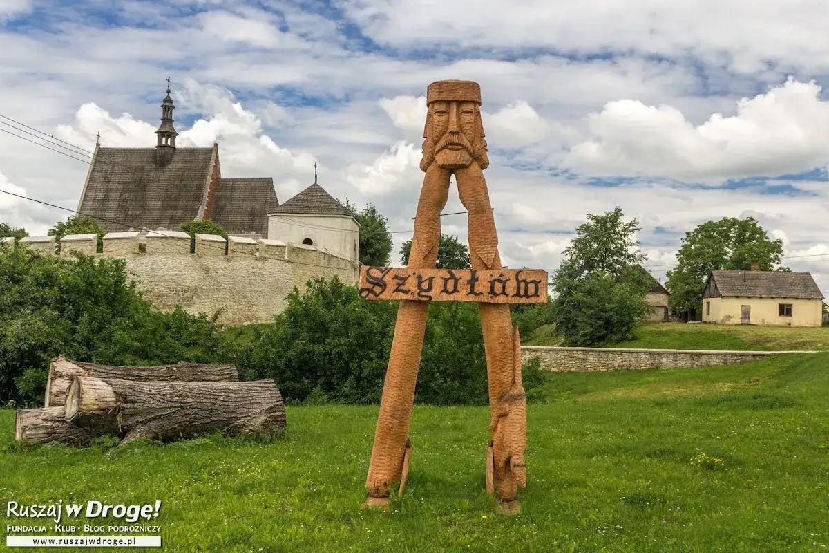 Drewniana rzeźba wita w Szydłowie, jednej z atrakcji G&oacute;r Świętokrzyskich. W tle zamek i wiejskie domki.