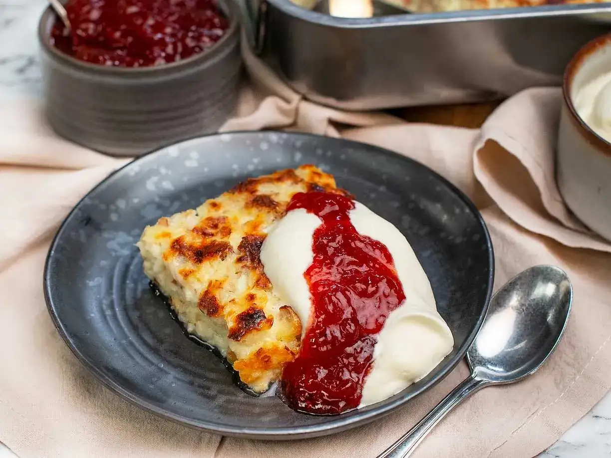 En bit gyllene ostkaka med keso, toppad med vispgrädde och rårörda lingon. Serveras på en mörk tallrik med en sked.