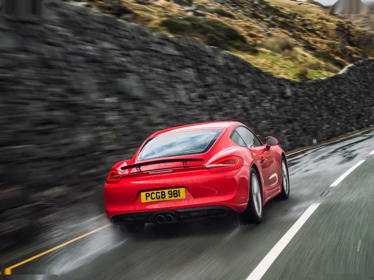 Czerwony Porsche Cayman mknie po mokrej drodze, rozpryskując wodę.