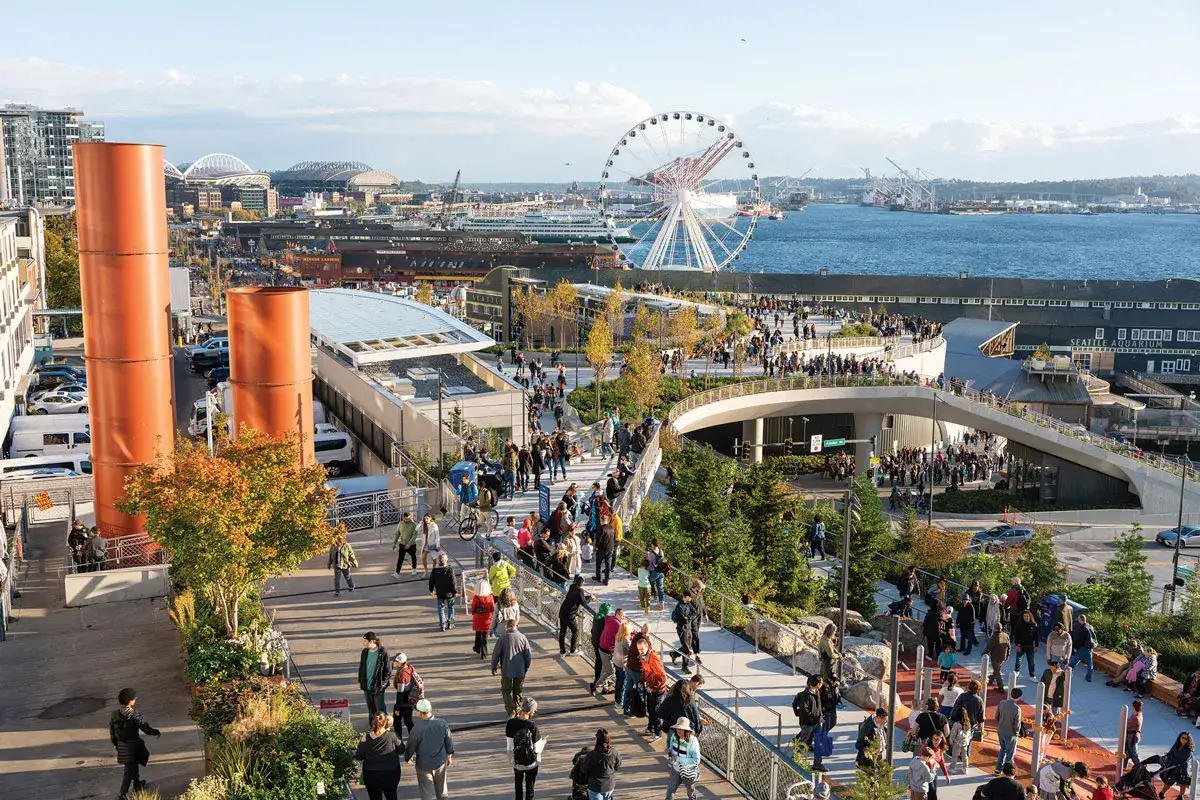 A vibrant scene of Seattle street photography, with people strolling along elevated walkways, the Great Wheel in the distance, and iconic orange silos framing the view.