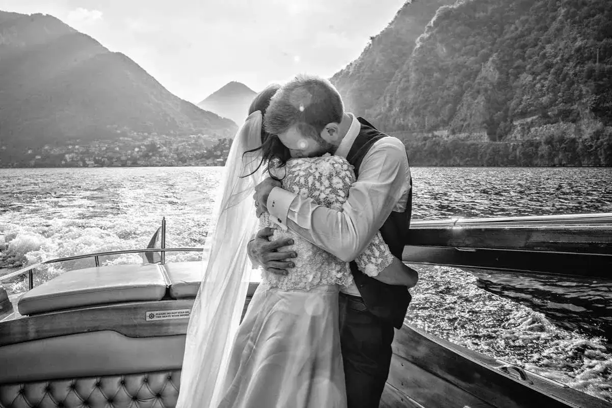 Un abbraccio intenso tra gli sposi su una barca, con montagne sullo sfondo. Dettagli matrimonio che catturano l'amore e la gioia del momento.