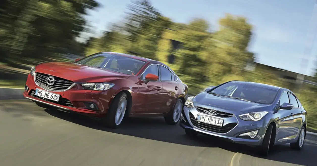 Czerwona Mazda 6 2025 i niebieski Hyundai jadą obok siebie po krętej drodze, otoczone rozmytym tłem drzew.