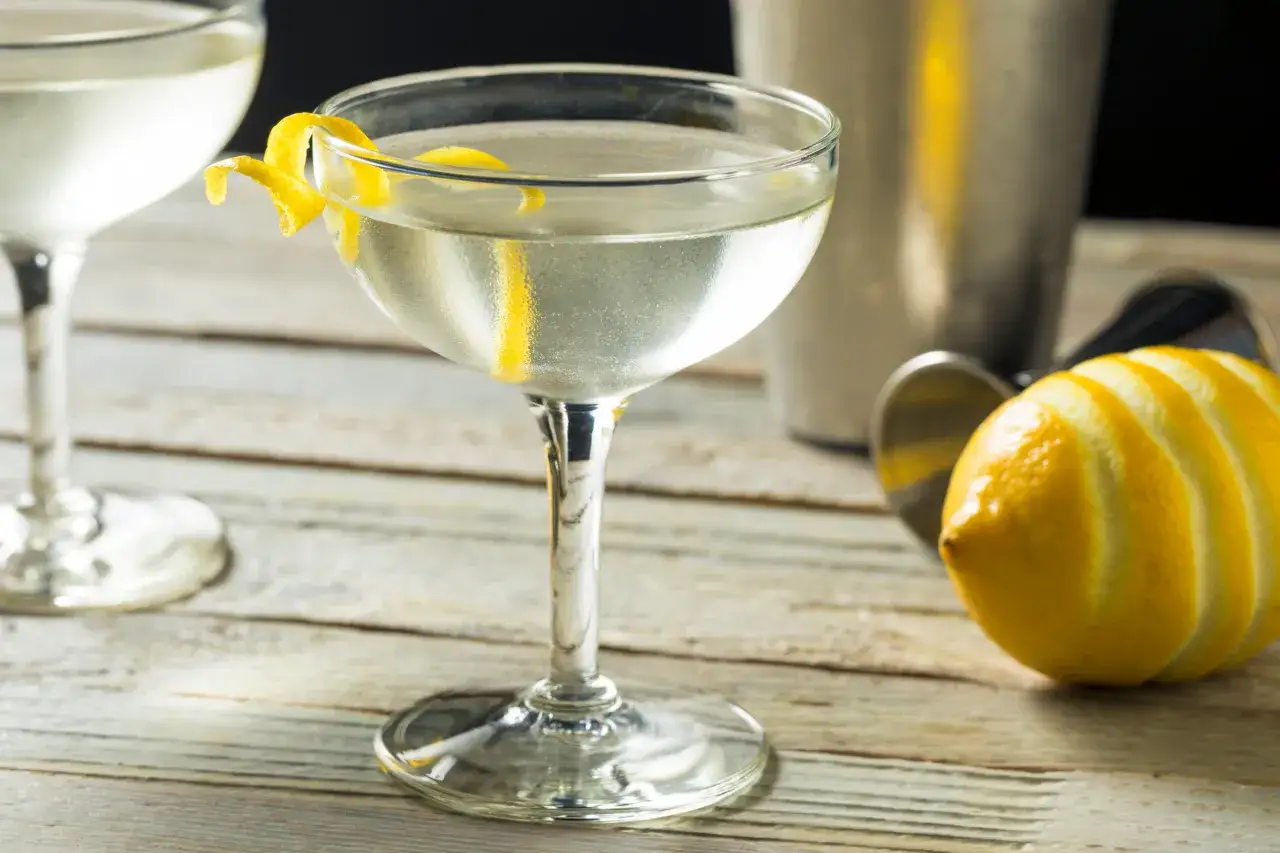 En elegant drink, som en james bond drink, serveras i ett coupeglas med en citronskal. En citron och en shaker syns i bakgrunden.