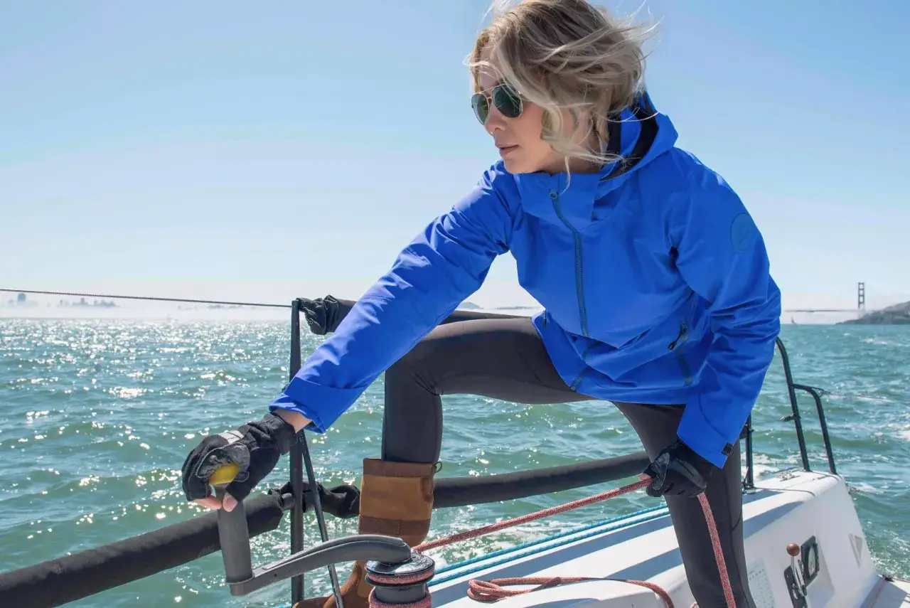 Donna con abbigliamento da barca a vela, occhiali da sole e giacca blu, manovra la barca a vela con la Golden Gate Bridge sullo sfondo.
