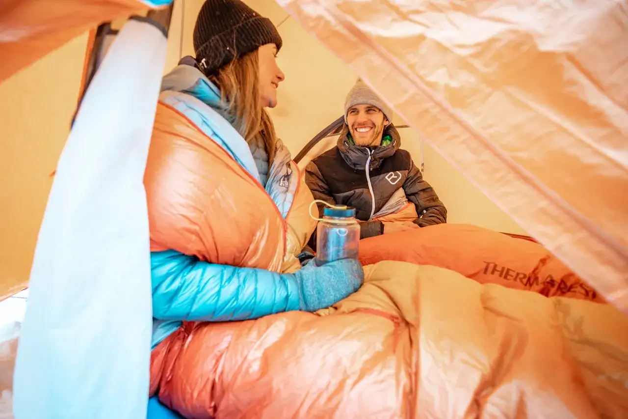 Couple souriant dans une tente, emmitouflés dans des sacs de couchage orange vif. Ils semblent prêts à bien dormir en tente, profitant de la chaleur et de la compagnie.