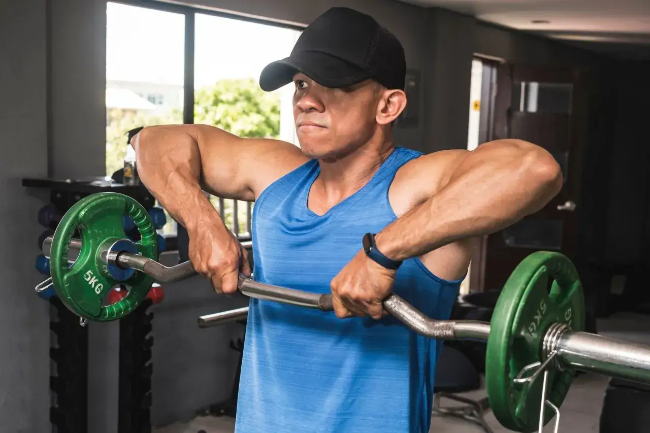 Man i blå linne och keps utför ett **drag till hakan** med en skivstång i ett gym.