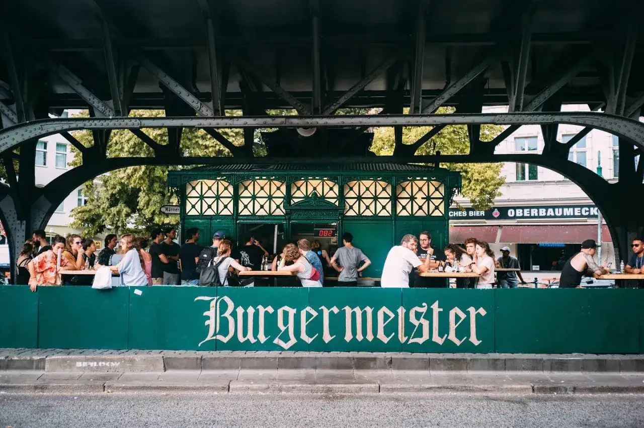Menschen genie&szlig;en Burger und Bier unter der U-Bahn-Br&uuml;cke. Das gr&uuml;ne Schild mit der Aufschrift 