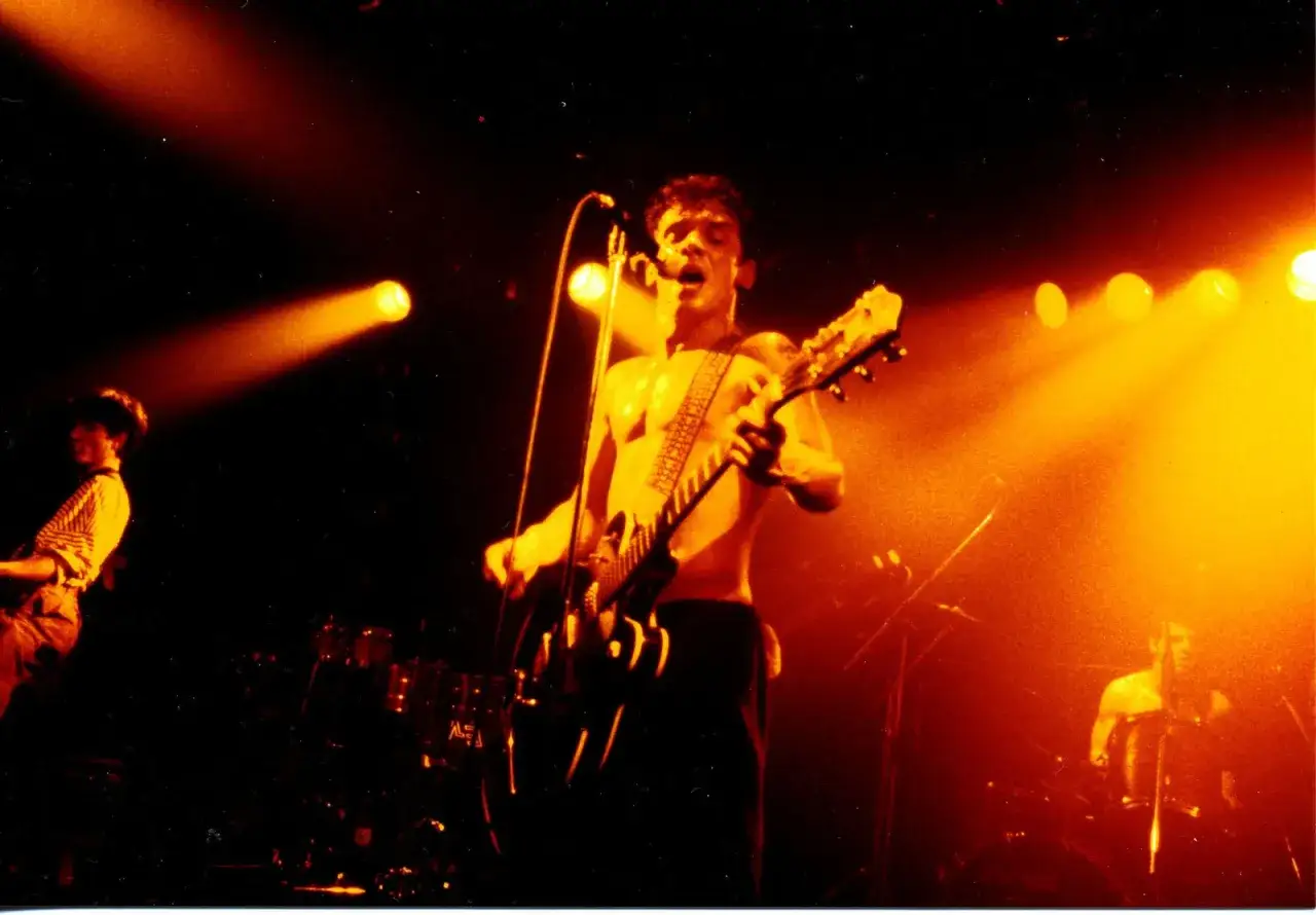 Un chanteur torse nu d'un groupe rock alternatif français hurle en jouant de la guitare sous des projecteurs orangés.