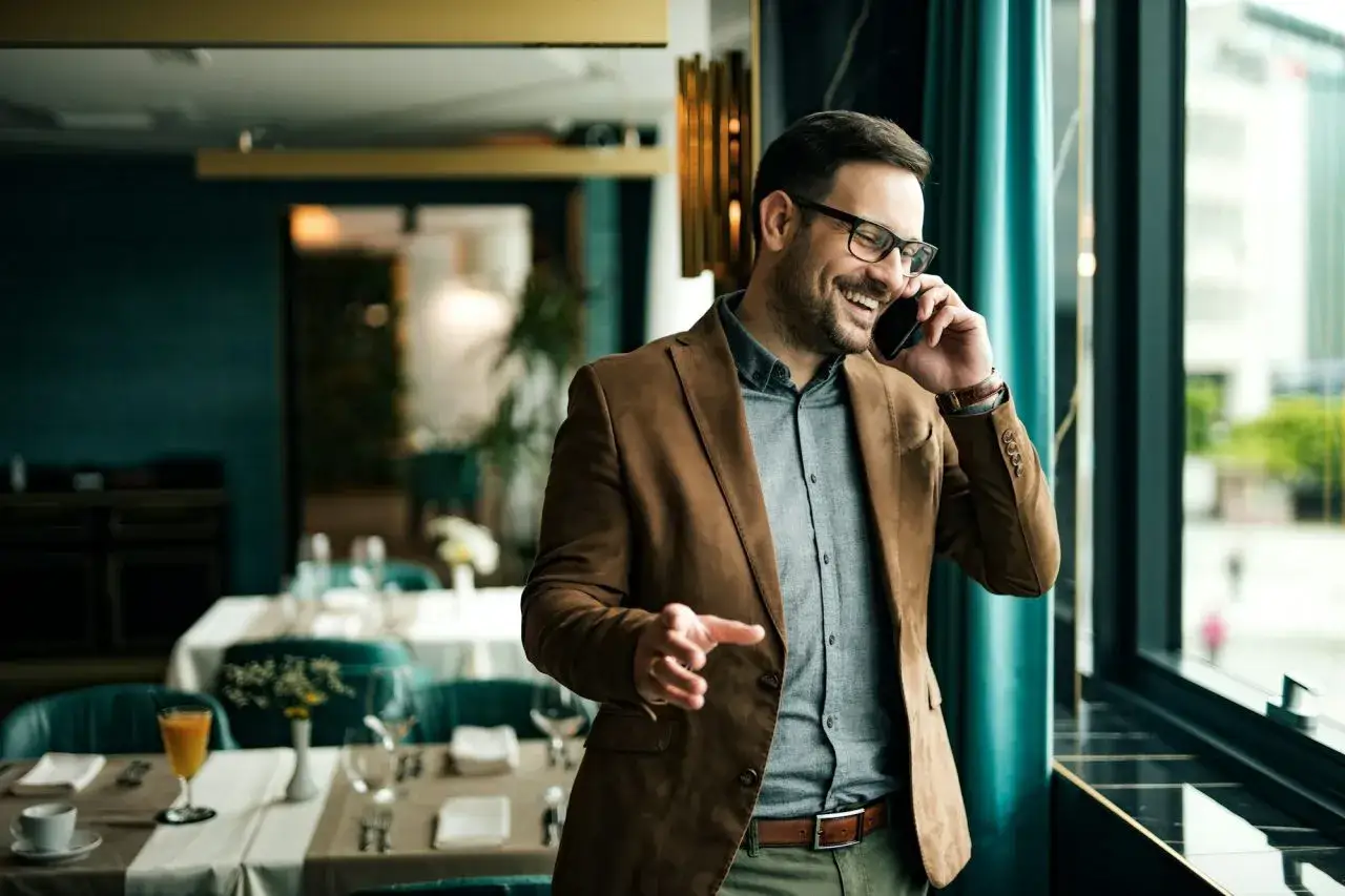 Uśmiechnięty mężczyzna w brązowej marynarce rozmawia przez telefon w eleganckiej restauracji.
