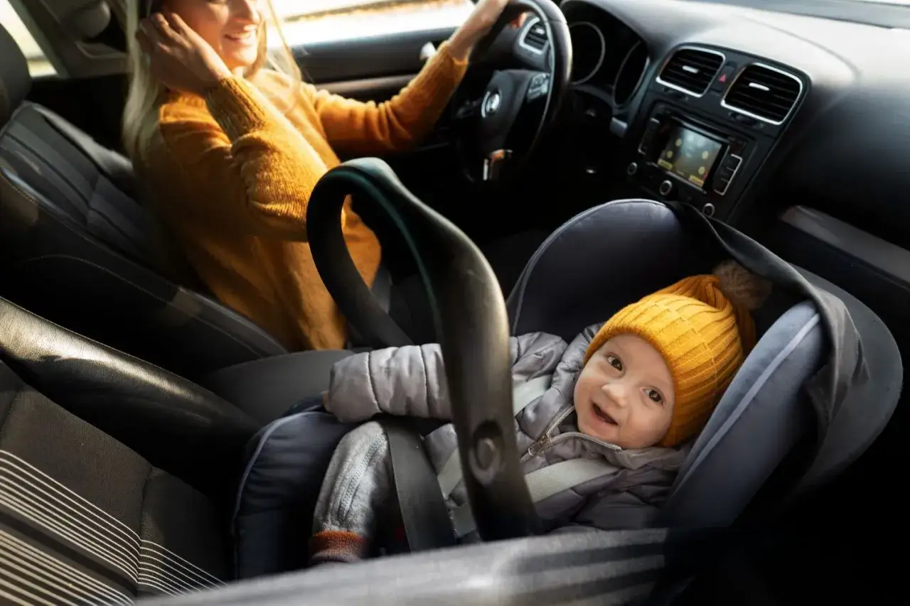 Uśmiechnięta kobieta prowadzi auto, a na przednim siedzeniu w foteliku podróżuje dziecko. Przewożenie dziecka na przednim siedzeniu jest dozwolone, gdy fotelik jest tyłem do kierunku jazdy i wyłączona jest poduszka powietrzna.