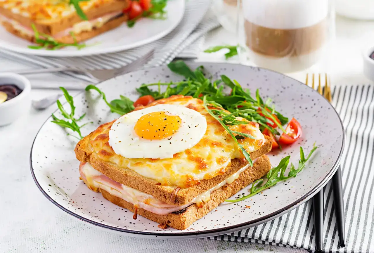 Ein k&ouml;stlicher Croque Madame mit geschmolzenem K&auml;se, Schinken und einem Spiegelei, serviert mit Rucola und Kirschtomaten.