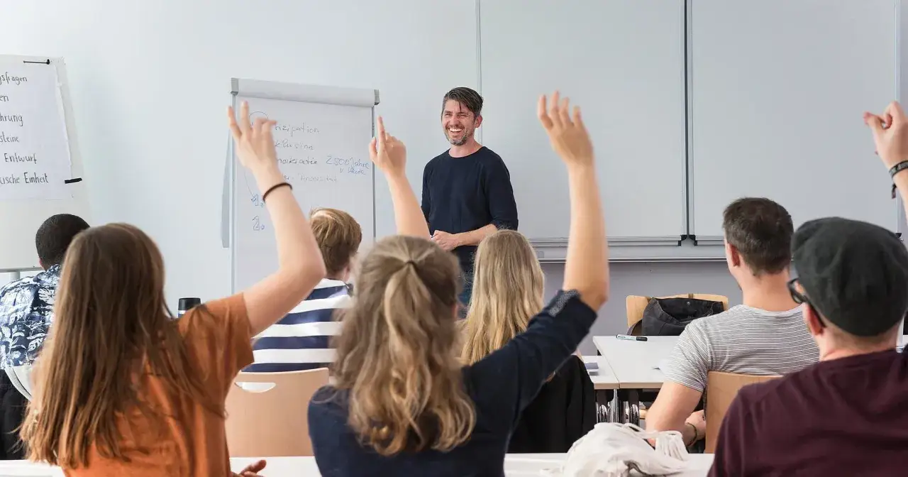 Ein Lehrer lacht, während Studierende mit erhobenen Händen Fragen stellen. Ein lebendiges Studium mit Menschen.