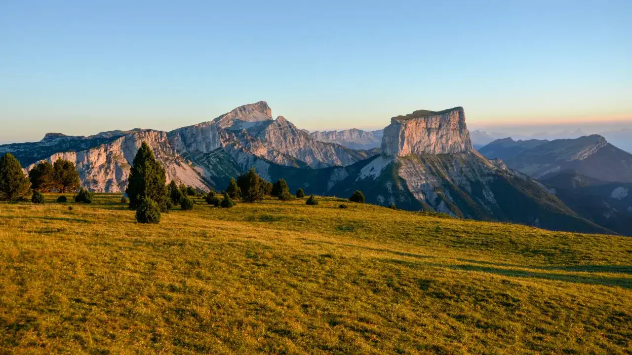 Sonnenlicht beleuchtet die Wiesen und den markanten Mont Aiguille. Gr&uuml;ne B&auml;ume stehen auf dem H&uuml;gel vor den zerkl&uuml;fteten Bergen.