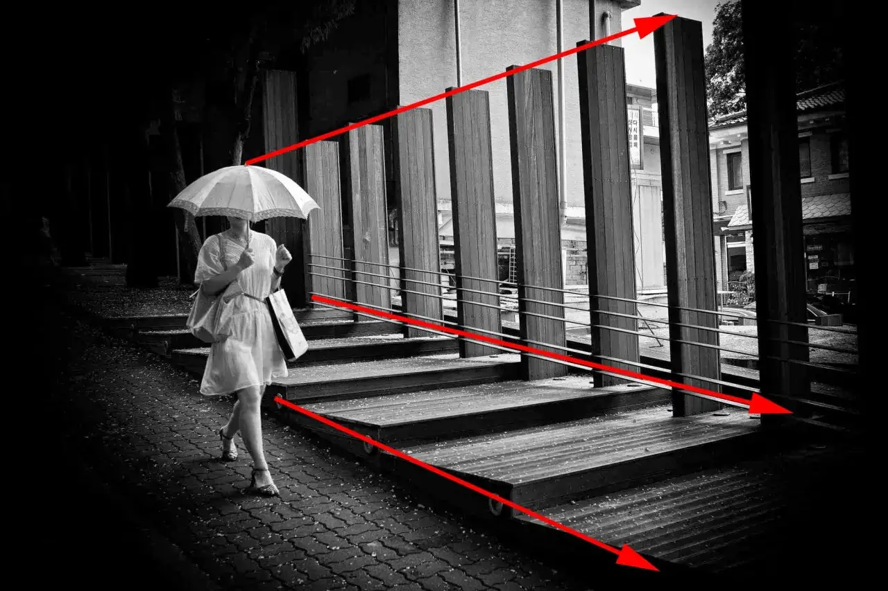 Kobieta z parasolką idzie po schodach. Czerwone linie wskazują kierunek i perspektywę, tworząc dynamiczną kompozycję.
