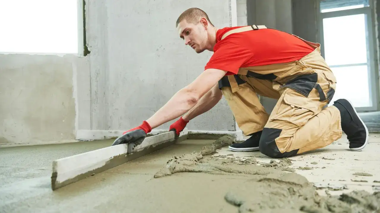 Budowlaniec w czerwonej koszulce i brązowych ogrodniczkach wyrównuje świeży, mokry beton na posadzce za pomocą łaty.