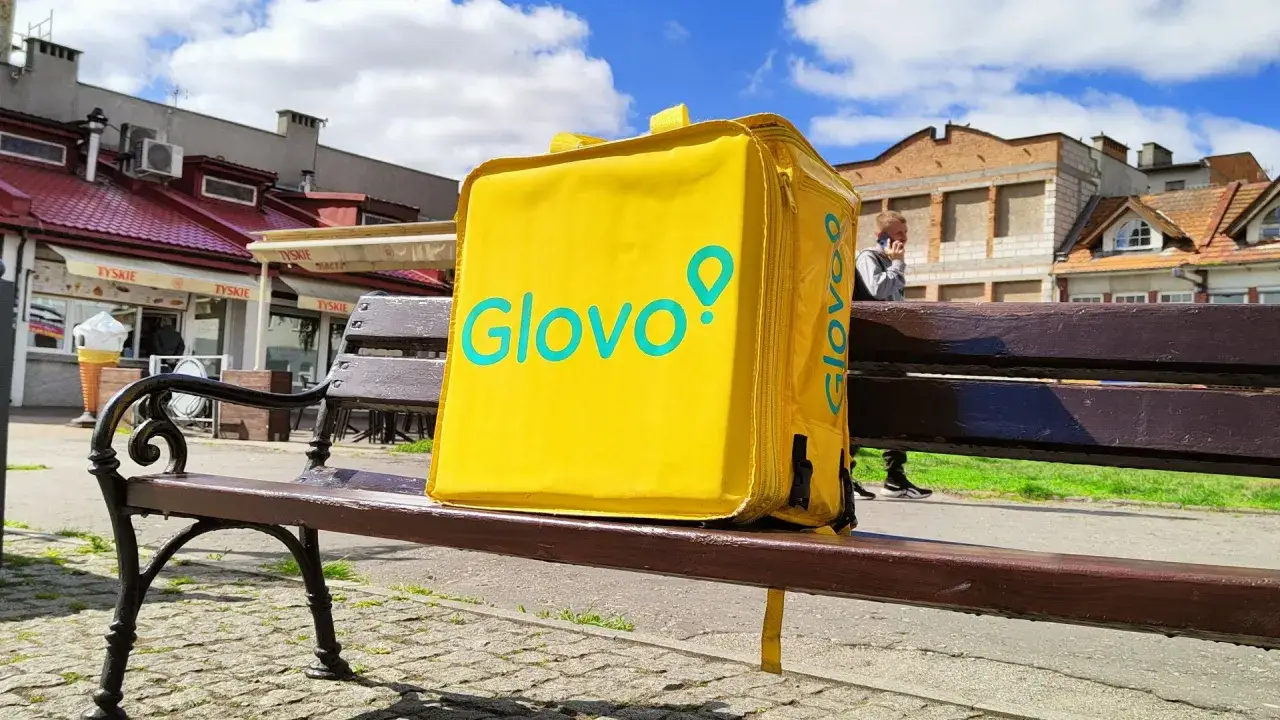 Ż&oacute;łta torba Glovo leży na ławce, czekając na kuriera pieszo. W tle widać budynki i błękitne niebo.