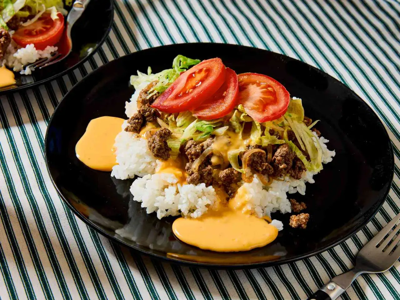 A plate of Okinawa taco rice, featuring white rice topped with seasoned ground beef, shredded lettuce, sliced tomatoes, and a drizzle of cheese sauce.