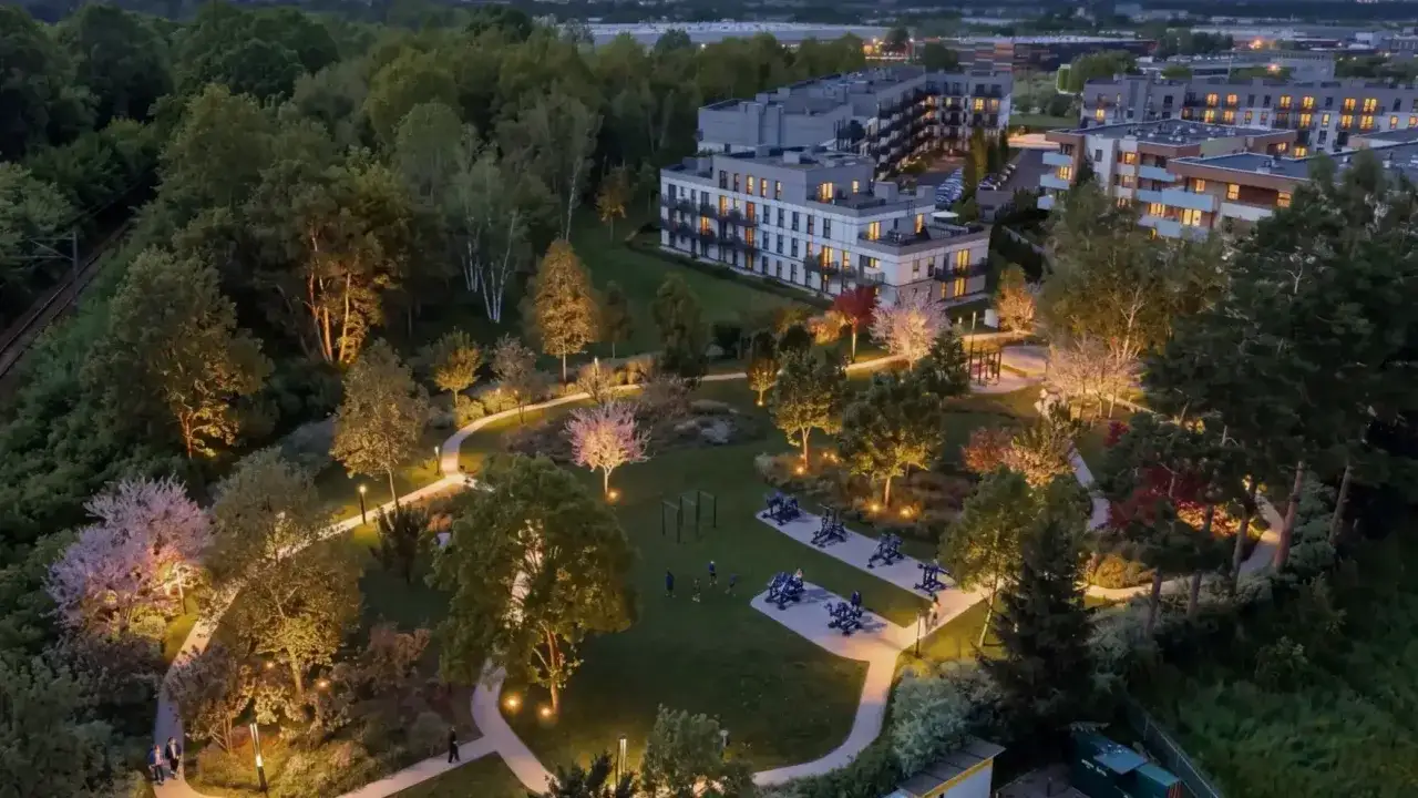 Oświetlony wieczorem centralny park z alejkami, drzewami i placem zabaw dla dorosłych, otoczony nowoczesnymi budynkami mieszkalnymi.