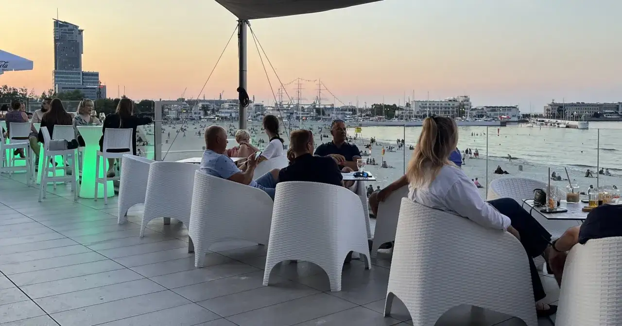 Wieczorny relaks w Gdyni. Ludzie siedzą przy stolikach na tarasie z widokiem na plażę i morze. To idealne gdynia atrakcje dla dorosłych.