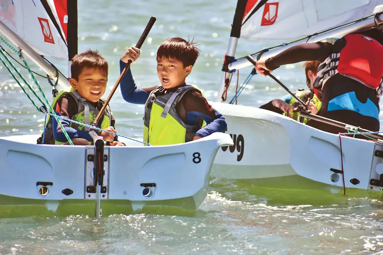 Deux enfants souriants apprennent la voile lors d'une colonie de vacances. Ils naviguent sur un petit bateau, le soleil brille sur l'eau.