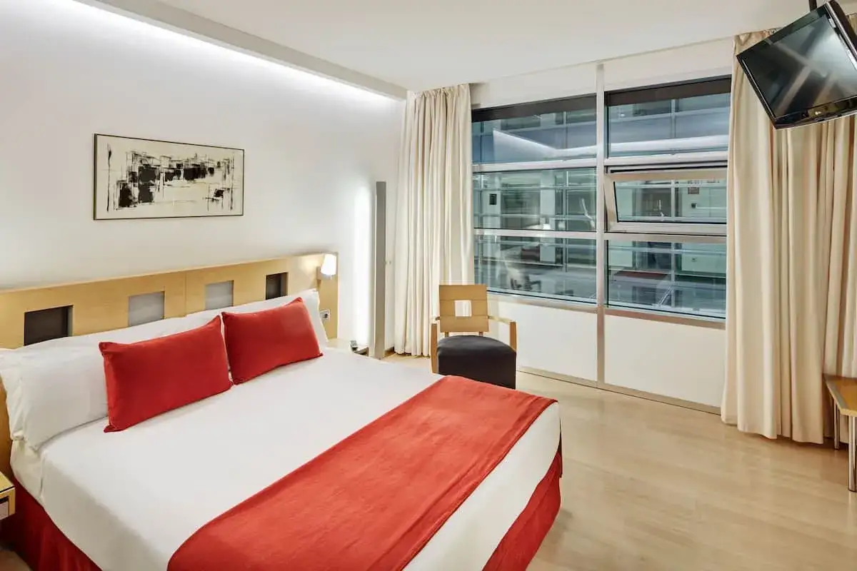 Habitación moderna en Sercotel Caspe con cama doble, cojines rojos, cuadro abstracto y gran ventanal con vistas.