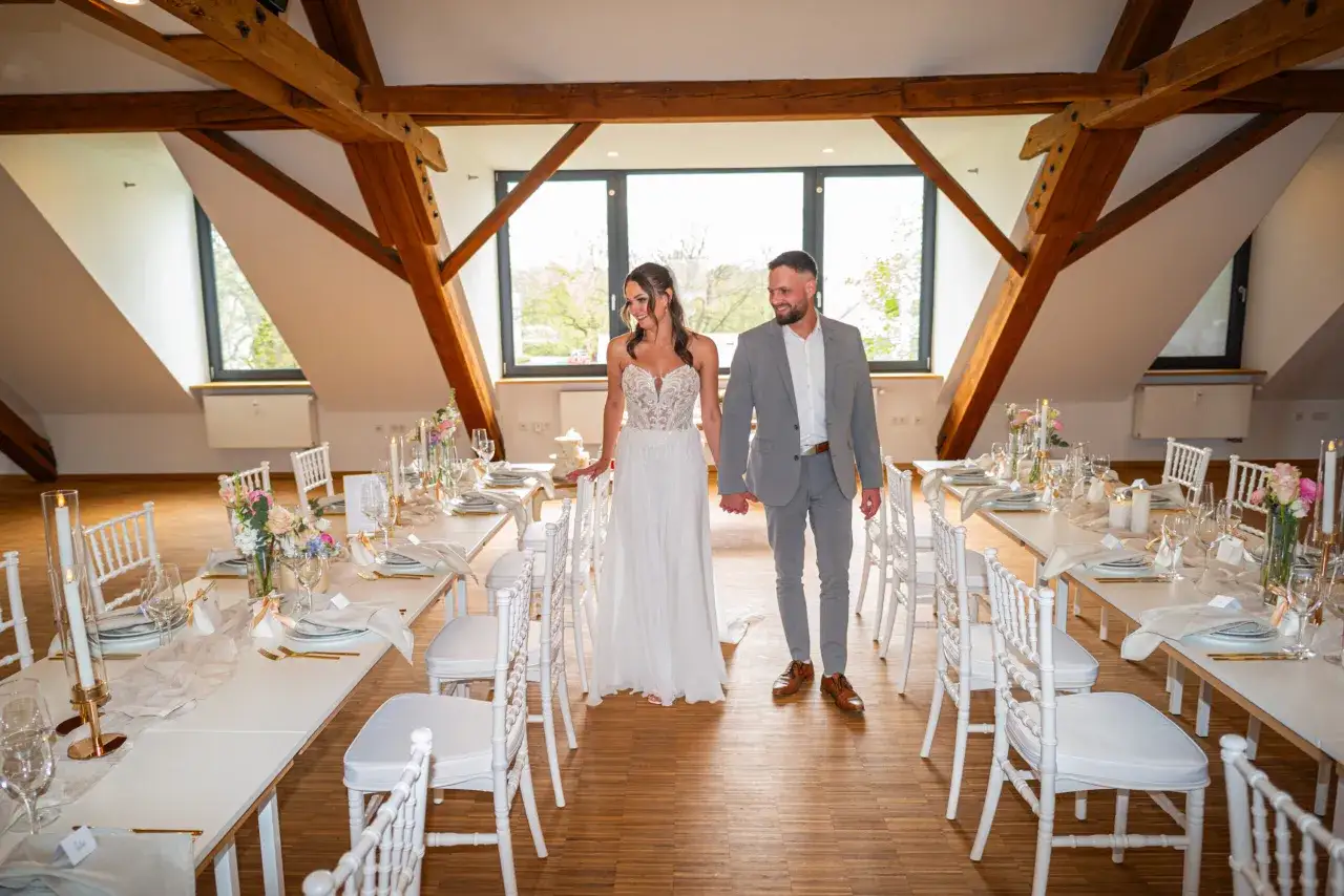 Brautpaar schreitet durch den Raum einer minimalistischen Hochzeit. Weiße Stühle und gedeckte Tische mit Kerzen und Blumen schaffen eine elegante Atmosphäre.