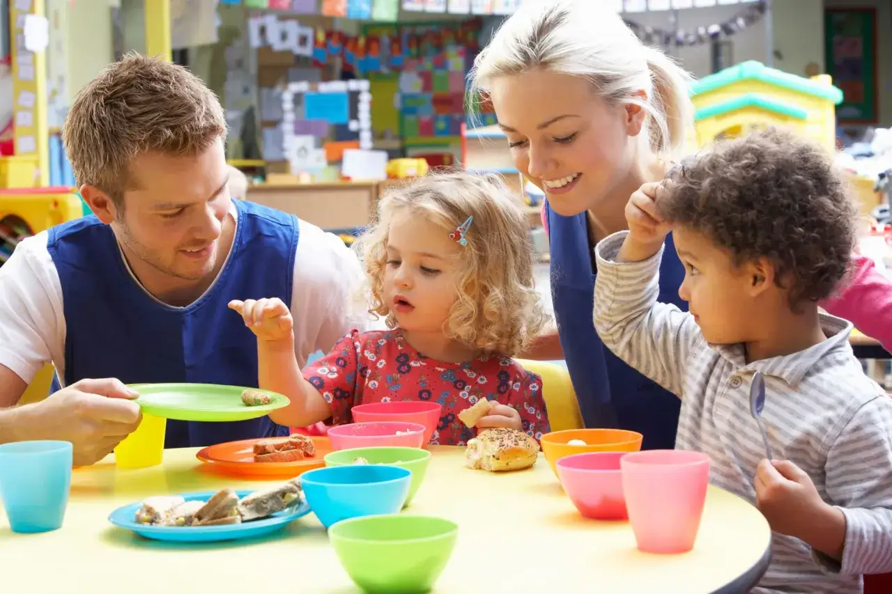 Un homme et une femme souriants, peut-être en formation de **master petite enfance à distance**, partagent un goûter avec deux enfants dans une salle colorée.