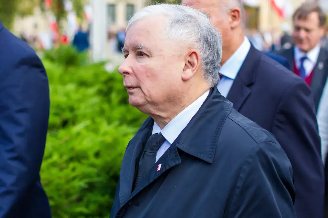 Jarosław Kaczyński w czarnym płaszczu, obok niego inni politycy. W tle zielone krzewy i budynki.