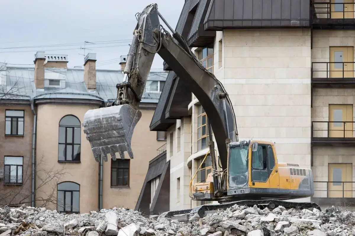 Koparka pracuje przy rozbiórce budynku, zasypując gruzem plac budowy.