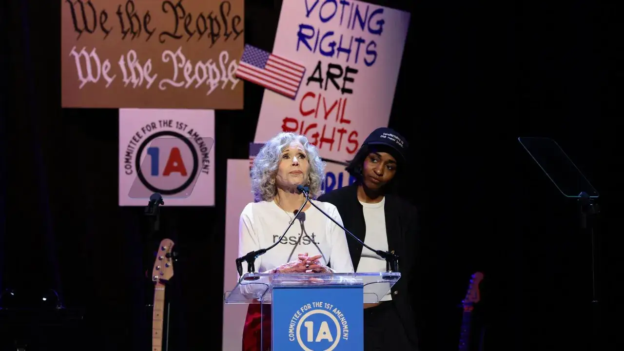 Jane Fonda spricht bei einer Veranstaltung, die an politische Musik erinnert. Hinter ihr stehen Banner mit Slogans wie 