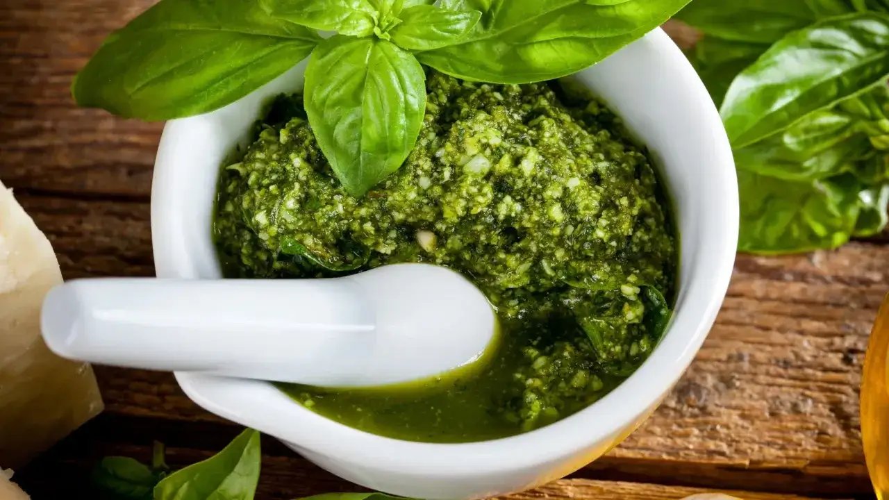 Świeże, aromatyczne pesto zielone w moździerzu, ozdobione listkami bazylii. Obok kawałek parmezanu i oliwa.