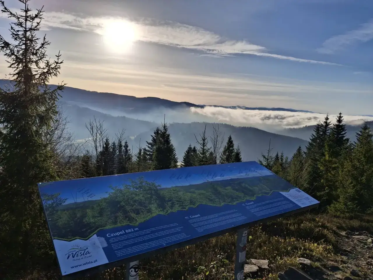 Widok z Czupla 882 m na pasmo górskie spowite mgłą. Tablica informacyjna przybliża panoramę.