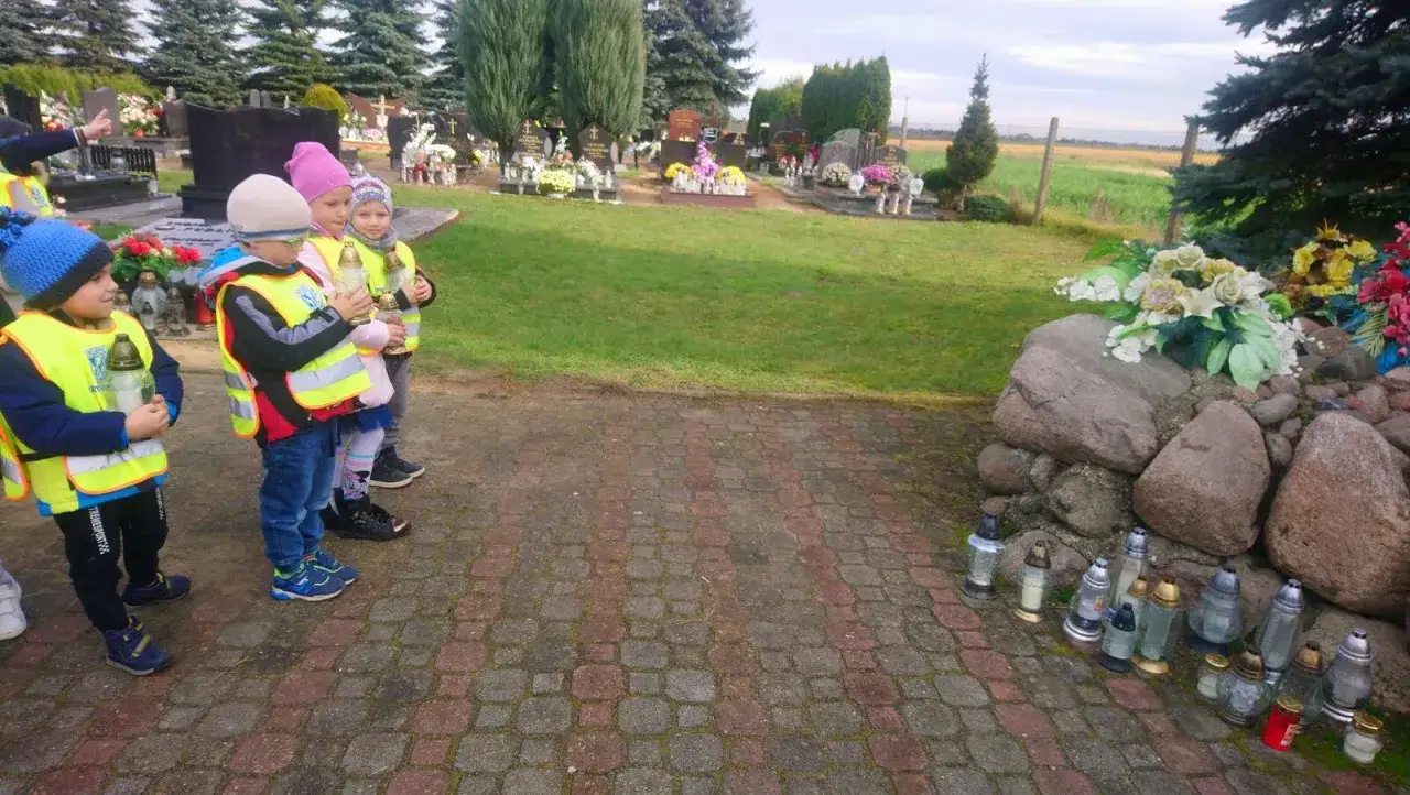 Dzieci w kamizelkach odblaskowych uczą się, jak przygotować się do pierwszej wizyty na cmentarzu z małym dzieckiem, trzymając znicze.