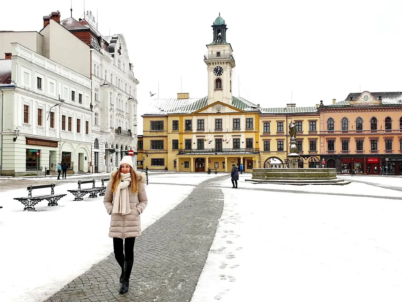 Kobieta w zimowym płaszczu spaceruje po ośnieżonym cieszyn rynek, z ratuszem i kolorowymi kamienicami w tle.