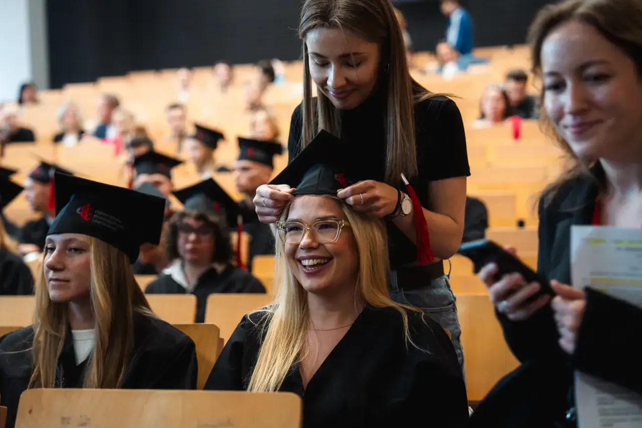 Uśmiechnięta studentka w okularach zakłada togę, gotowa na obronę magisterki w rok. W tle inni absolwenci w biretach.